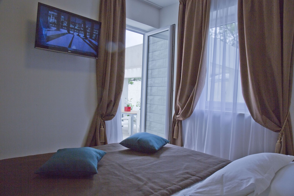 A bedroom with a bed, a TV mounted on the wall, and a view of the outdoors through a window.