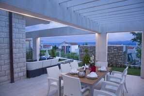 Un espacio de comedor al aire libre con pared de piedra, mobiliario de mimbre blanco y vistas a casas en el fondo.