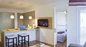 Ein modernes Hotelzimmer mit Küchenzeile, einem an der Wand montierten Fernseher, einem Bett mit weißer Bettdecke und einem Balkon mit Geländer.
