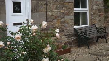 A stone building with a white door and window, a bench, and blooming roses.