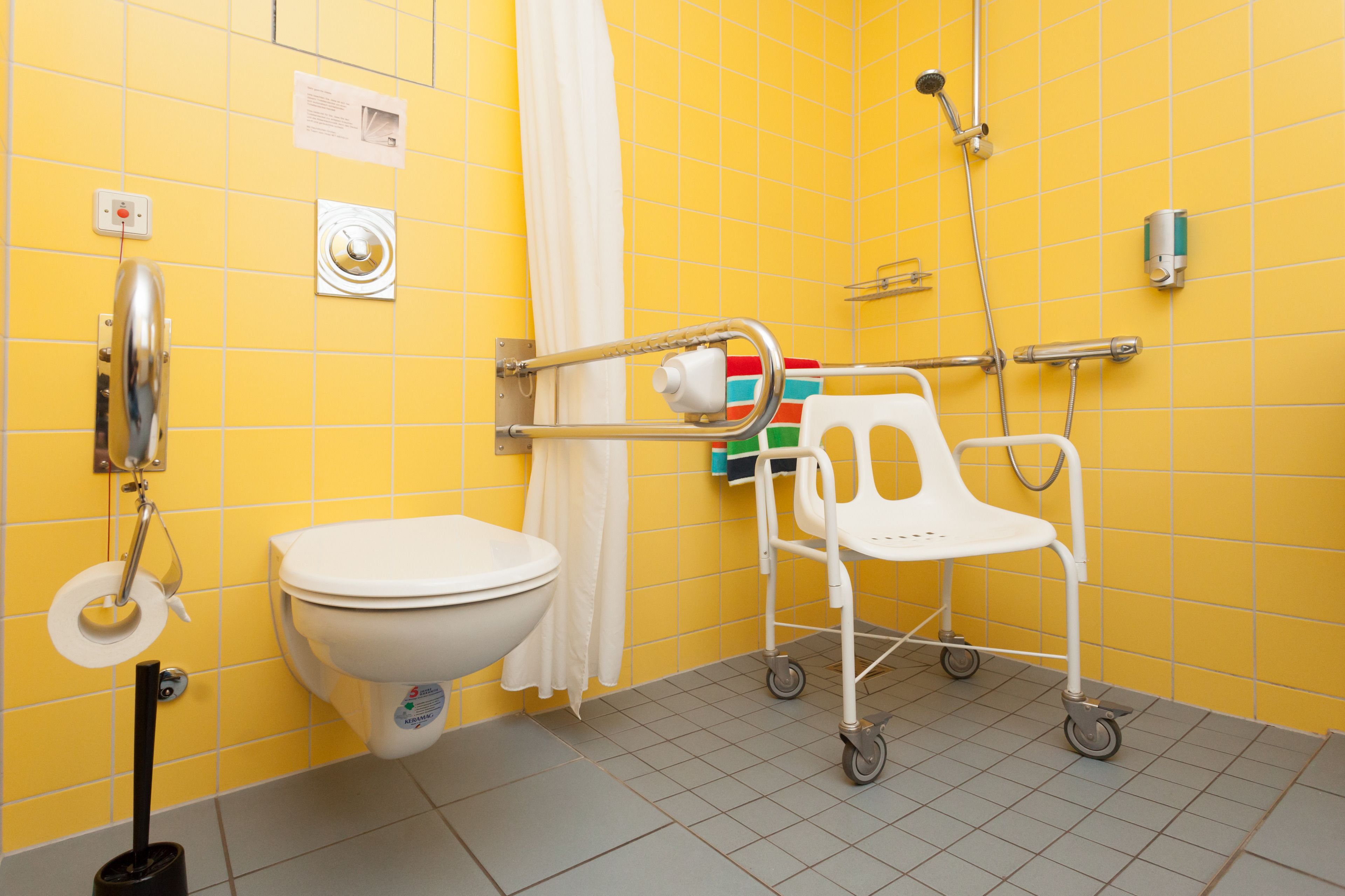 Un baño con azulejos amarillos, un inodoro blanco, una ducha y una silla de ruedas blanca.