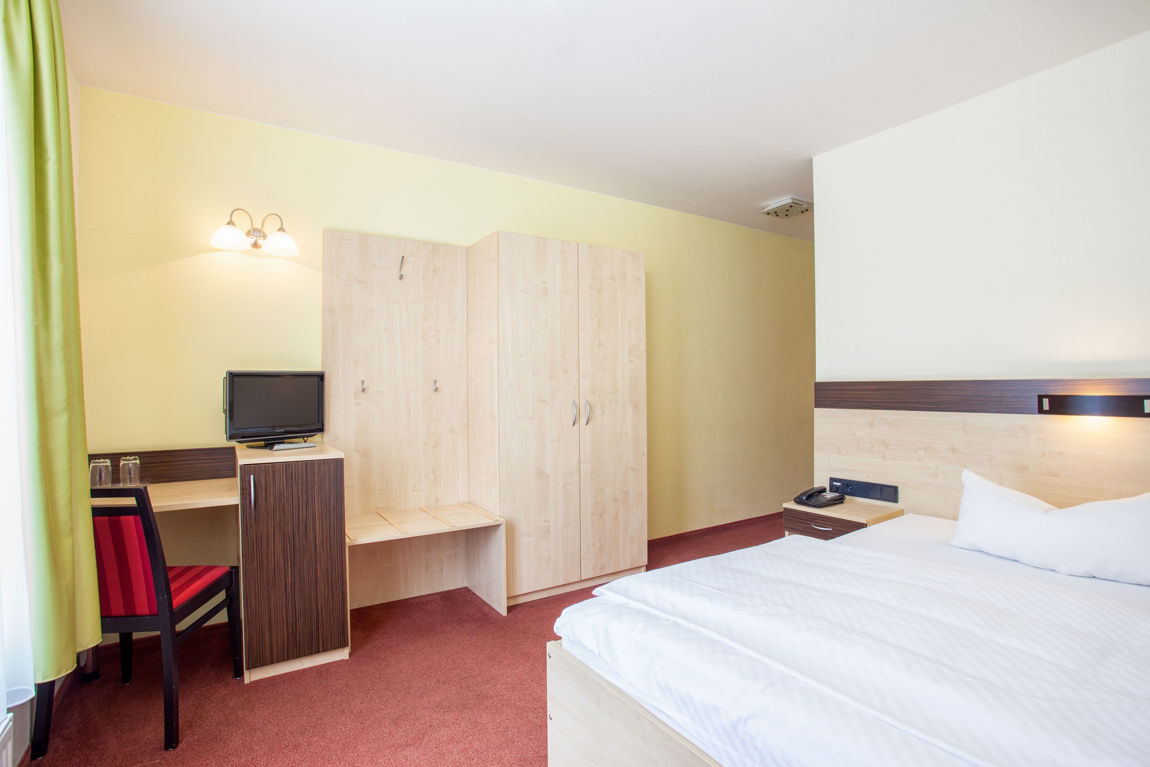 A hotel room with a bed, a desk with a chair, a television, and a wardrobe.