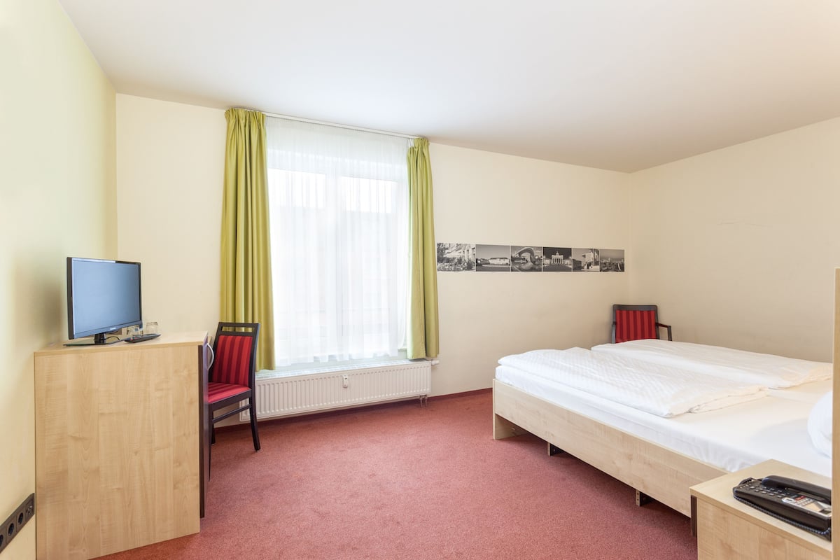 A hotel room with a bed, a desk with a computer, a chair, a radiator, and a window with curtains.