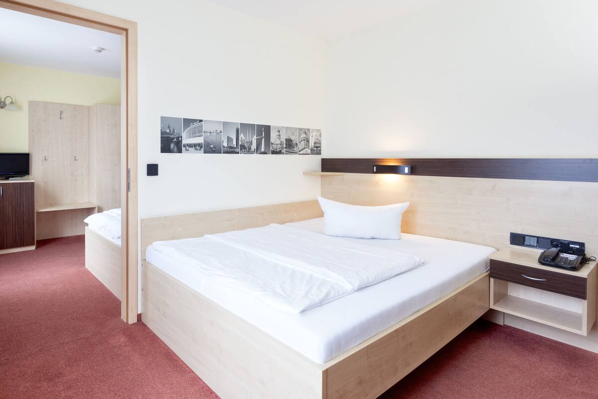 A hotel room with a wooden bed, a bedside table with a phone, a small desk with a chair, a television, and a wall-mounted light.