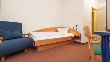 Una cama individual con cabecera de madera, un sillón azul, una mesita de noche con lámpara, y una silla con cojín verde.