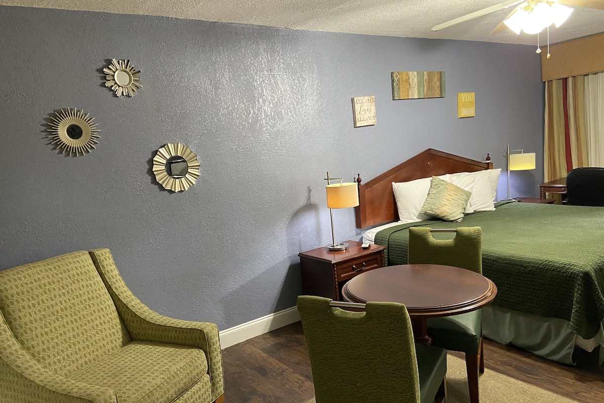A hotel room with a bed, a green armchair, a round table, and a nightstand with a lamp.