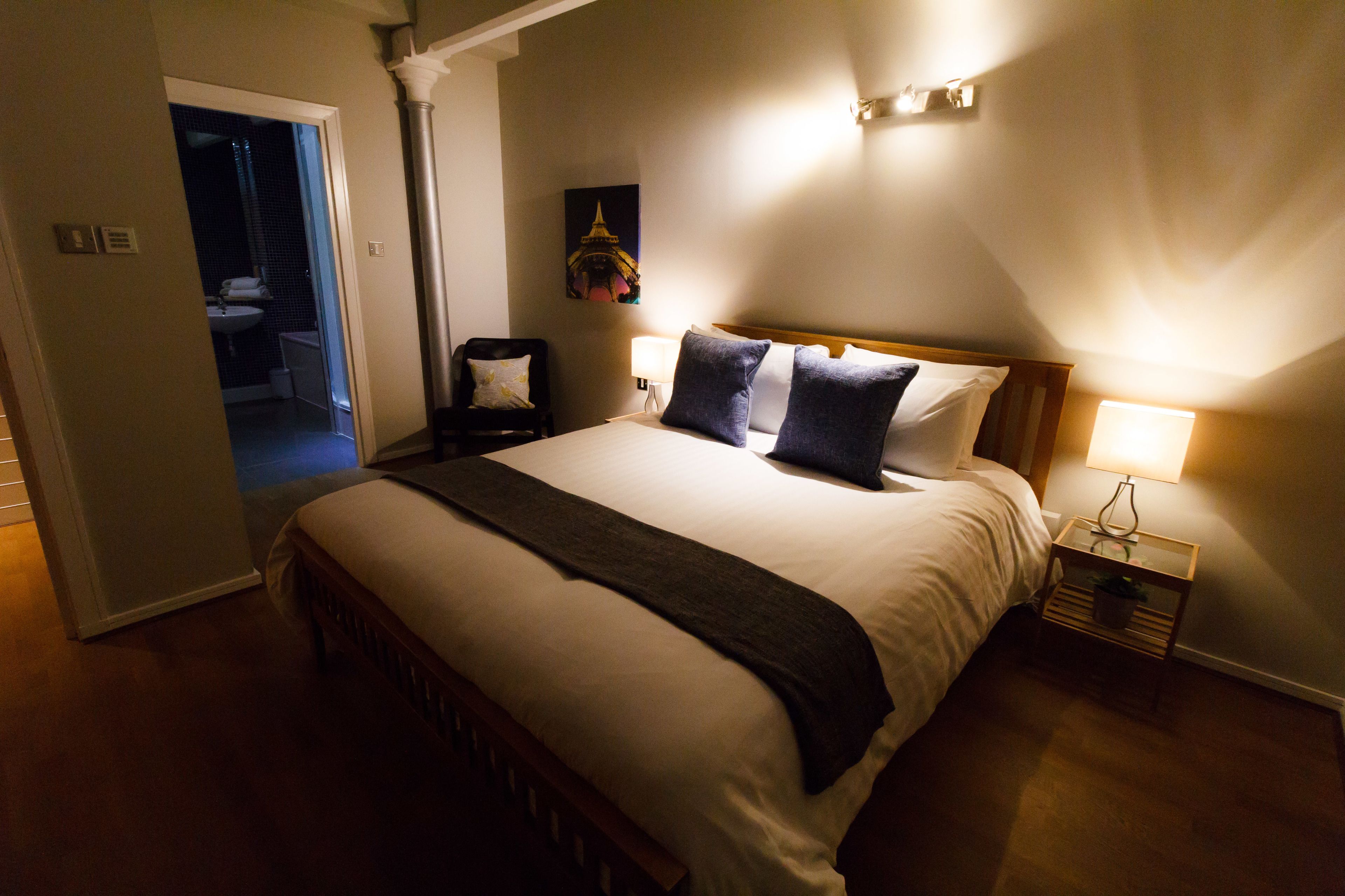 A hotel room with a large bed, a nightstand with a lamp, a chair, and a view of the bathroom.
