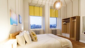 Ein Schlafzimmer mit Bett, zwei Fenstern, einem Kleiderschrank und Blick auf die Stadt.
