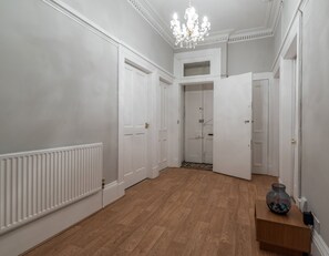 Ein Korridor mit Holzboden, weißen Wänden und einem Chandelier. Auf beiden Seiten befinden sich weiße Türen, und rechts steht ein kleiner Tisch mit einer Vase.