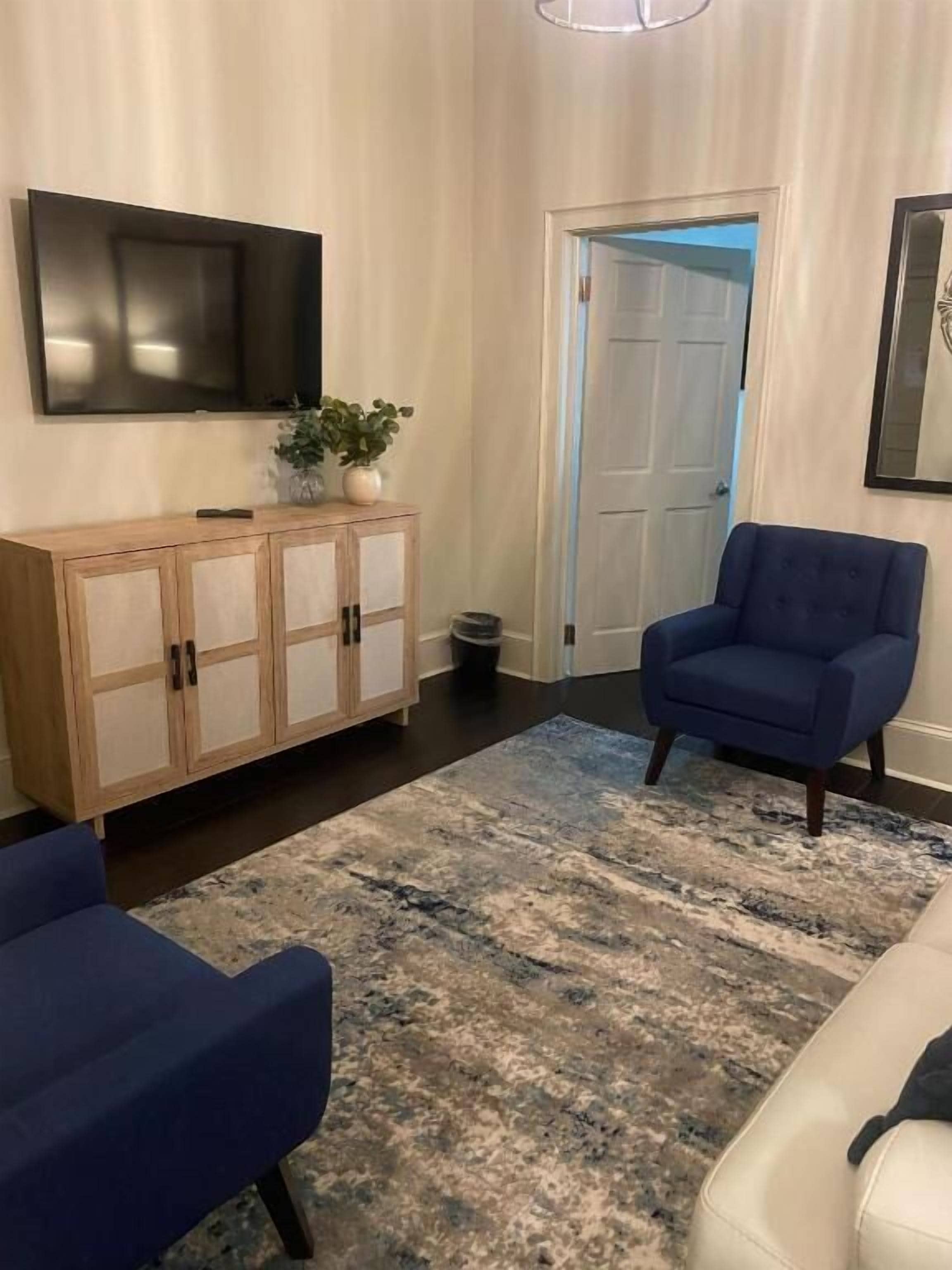 A living room with a blue sofa, a white sofa, a blue armchair, a wooden cabinet, and a large TV on the wall.