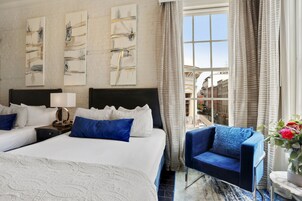 Quarto de hotel com uma cama grande, duas mesas de cabeceira, uma poltrona azul e vista para a cidade por uma janela ampla.