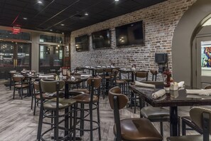 A restaurant with brick walls, multiple tables and chairs, and a television mounted on the wall. - La Galerie French Quarter Hotel (New Orleans)