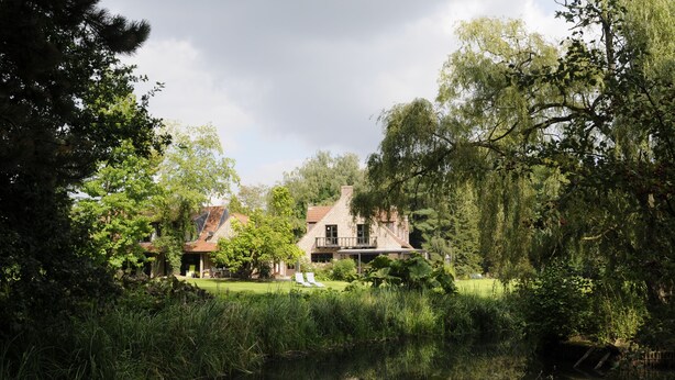 A serene lakeside setting with a traditional house featuring a stone facade and a red roof, surrounded by lush greenery and a calm water body. - 't Vijverhof (Geraardsbergen)