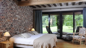 A bedroom with a brick wall, a large bed, a wooden nightstand, a chair, and a view of a garden through a glass door. - 't Vijverhof (Geraardsbergen)