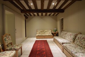 A spacious room with a high ceiling, wooden beams, a floral sofa, a matching armchair, and a red patterned rug.