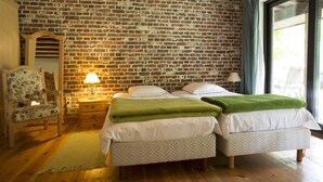 A room with a brick wall, two beds with white bedding, a green throw blanket, a wooden chair, a lamp, and a window with curtains. - 't Vijverhof (Geraardsbergen)