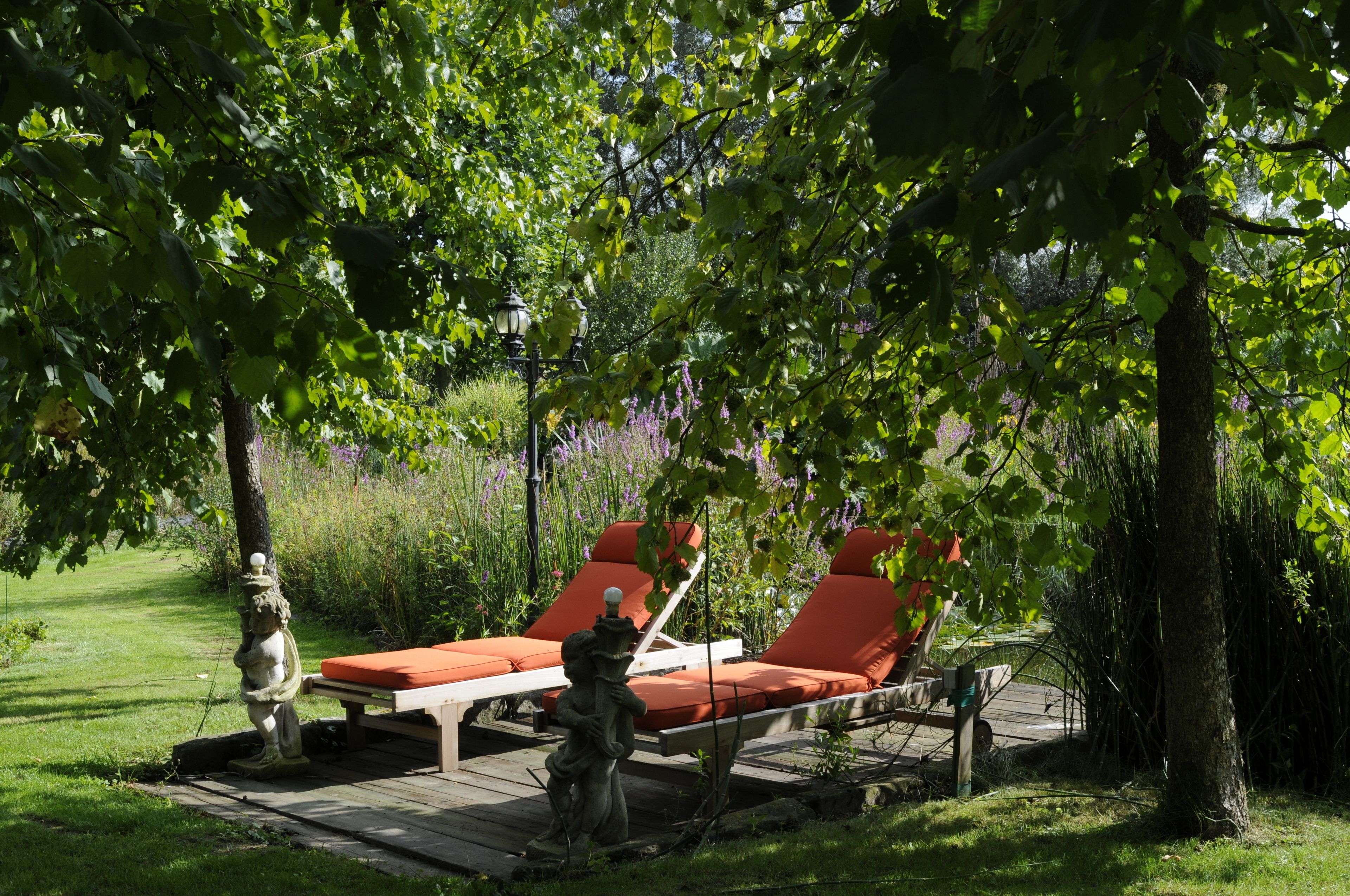 A garden with two red lounge chairs under a tree, a wooden deck, and a statue.