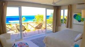 A bedroom with a large bed, a chair, a small stool, and a view of the ocean through large sliding doors. - Villa Encantada by Coco B Isla (Isla Mujeres)