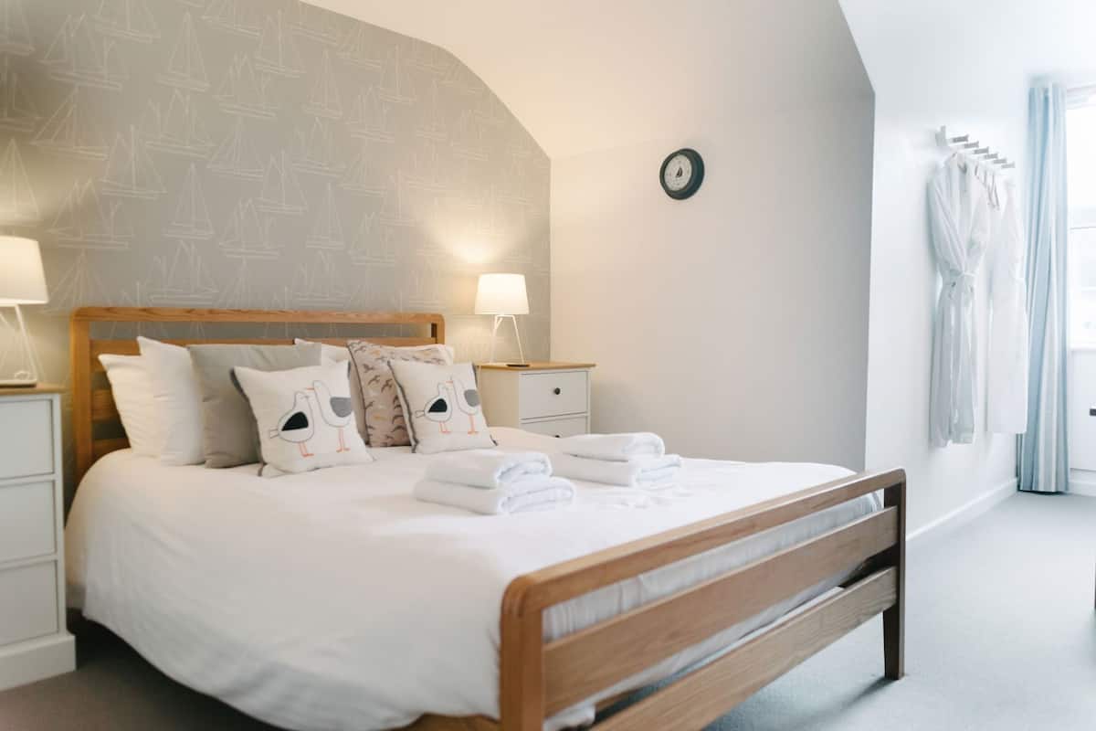A neatly made bed with pillows, a bedside table with a lamp, a clock on the wall, and a window with curtains.