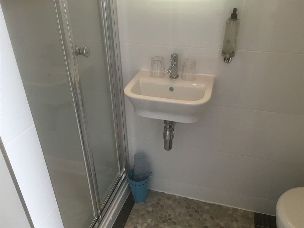 A bathroom with a glass shower enclosure, a sink with two clear glasses, a blue trash bin, and a toilet.
