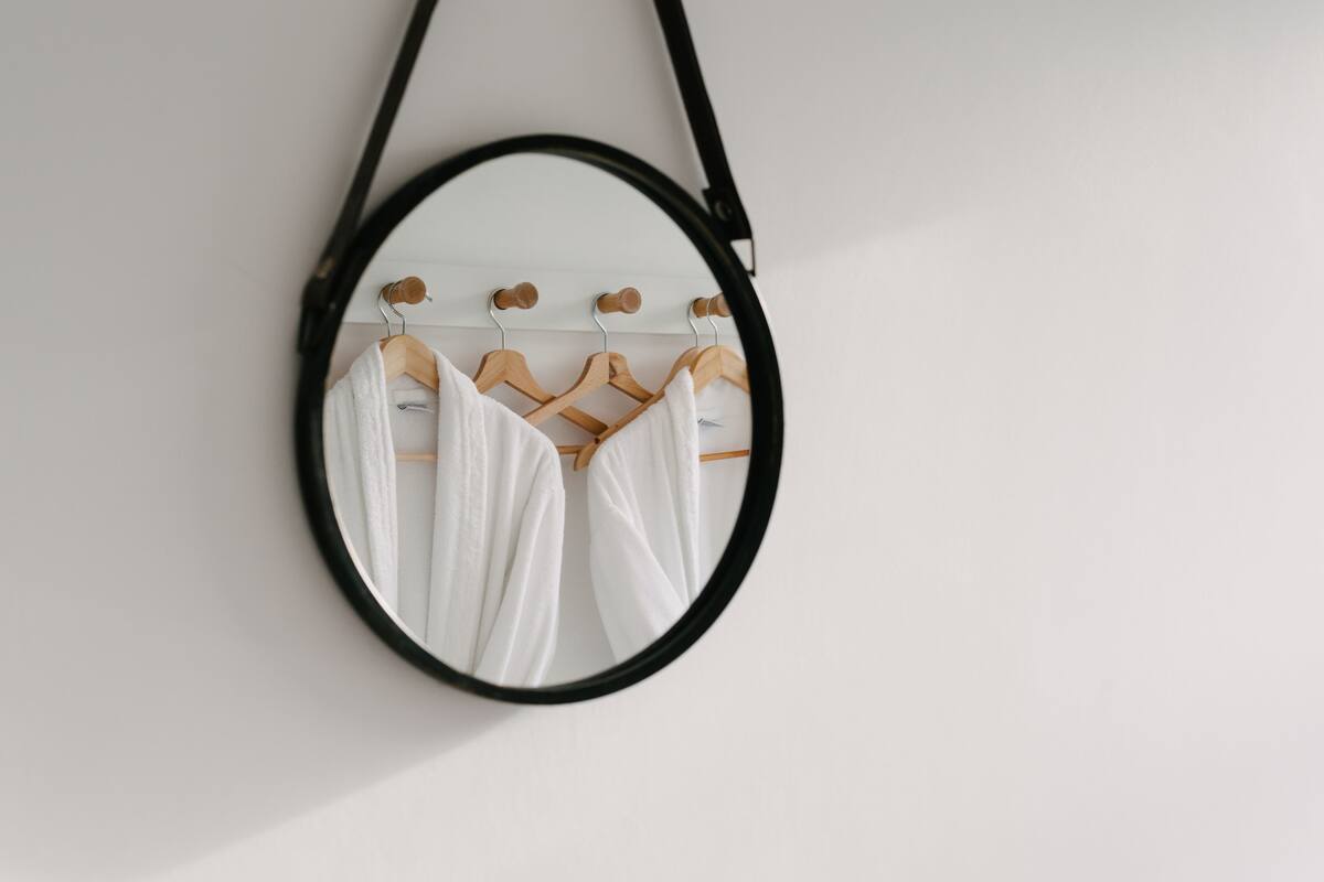 A mirror with a black frame reflecting a white wall and a wooden hanger with a white garment.