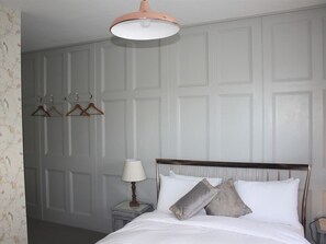 A neatly made bed with a wooden headboard, a bedside lamp, and three wooden hangers on a wall-mounted rack. - Portreath Arms (Redruth)