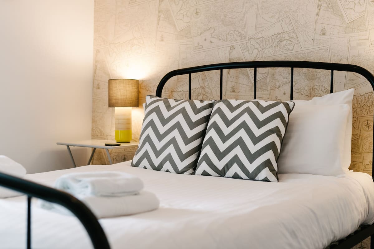 A neatly made bed with two patterned pillows, a bedside lamp, and a small table with a yellow object.
