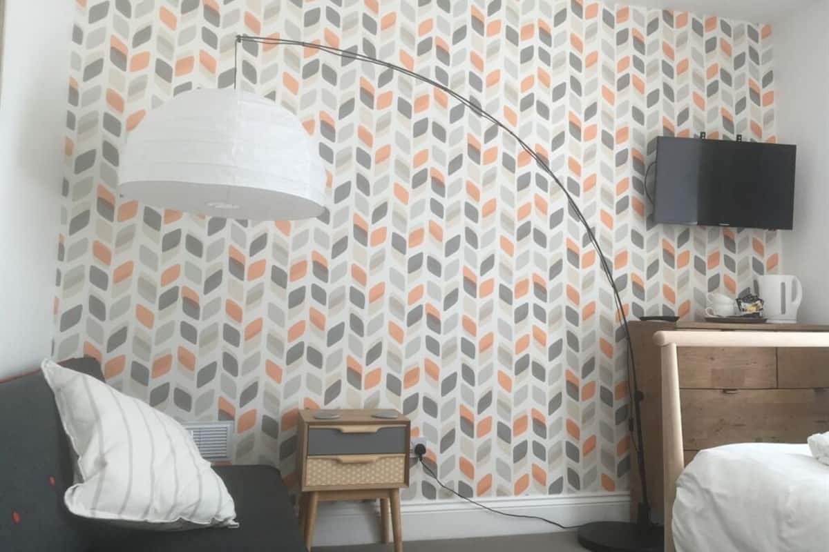 A room with a patterned wallpaper, a desk with a flat-screen TV, a lamp, and a bed.