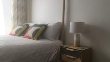 Una cama bien hecha con una colcha blanca y varias almohadas, una mesita de noche con lámpara y un pequeño aparador de madera con cajones.