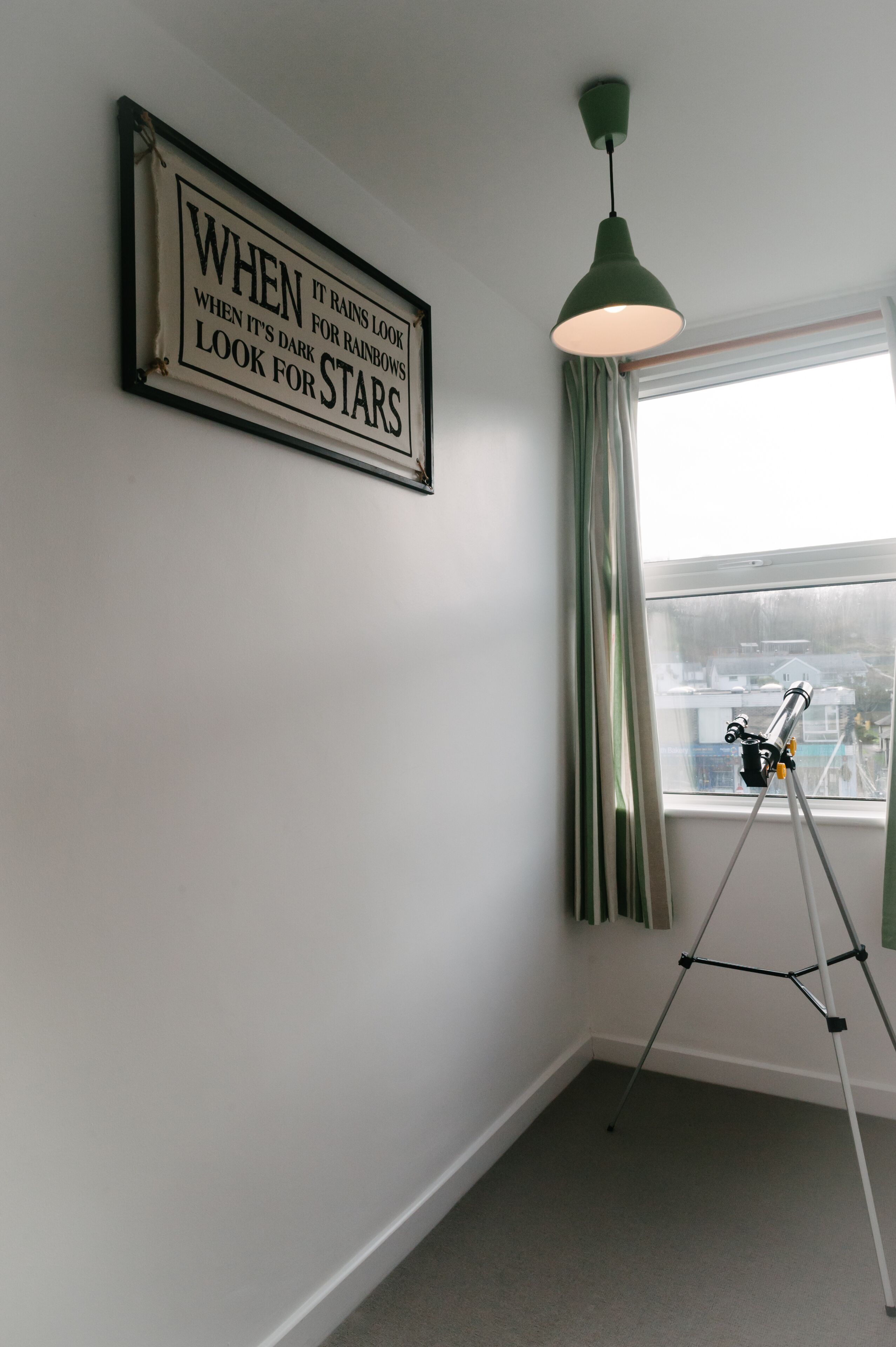 Een kamer met een ingelijst inspirerend citaat aan de muur, een telescoop op een statief, en een raam met gordijnen.