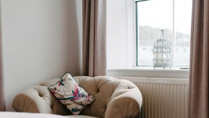 A corner of a room with a tufted armchair, a floral pillow, and a glass birdcage on a windowsill. - Portreath Arms (Redruth)