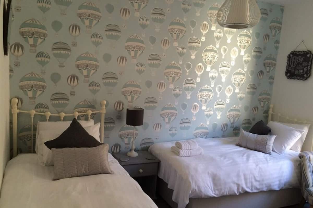 A bedroom with two beds, a nightstand, a lamp, and a wall with a pattern of hot air balloons.