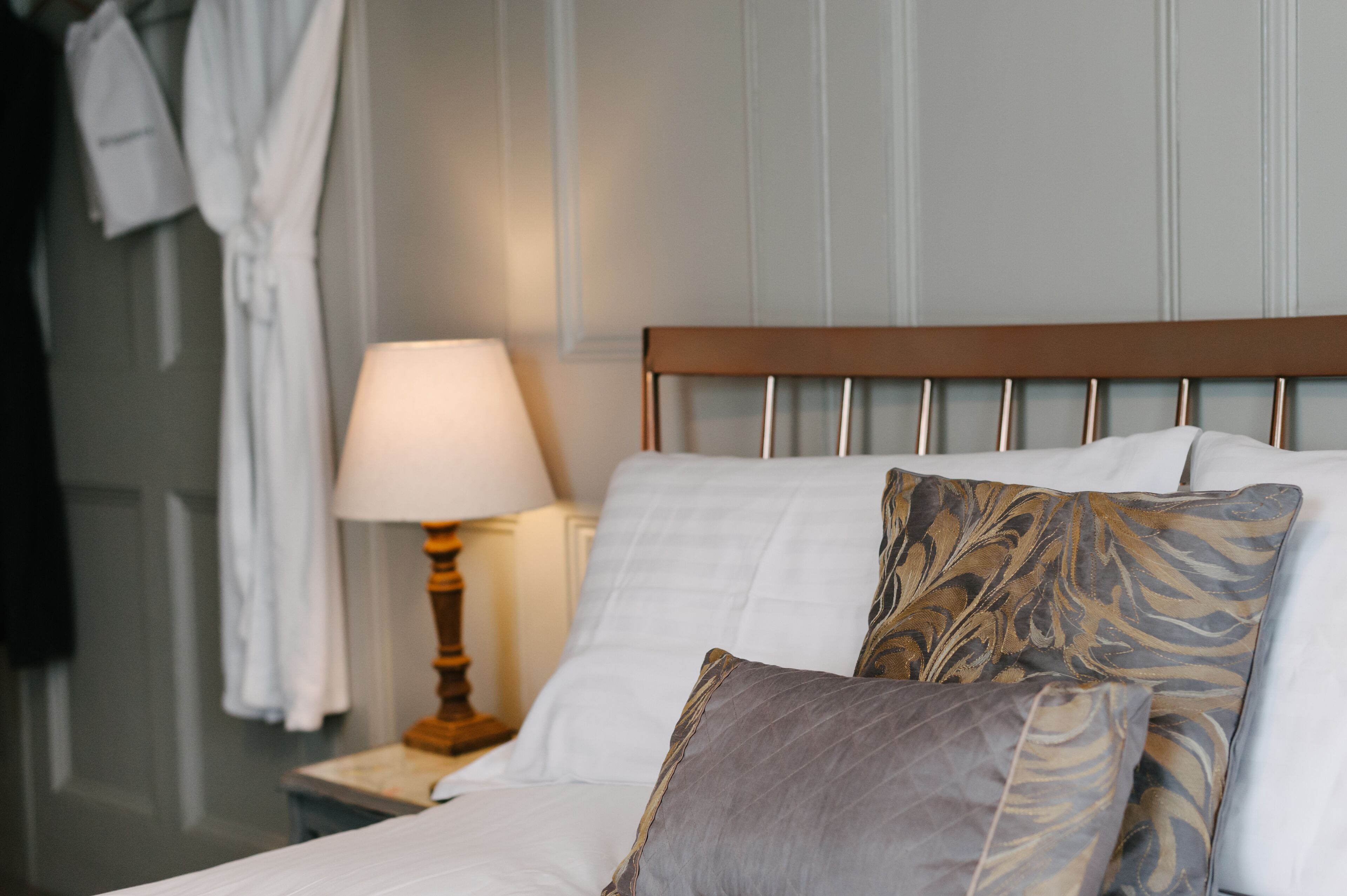 Un lit bien fait, avec une tête de lit en bois, une literie blanche et des coussins à motifs. On aperçoit une lampe de chevet et une table de chevet avec une autre lampe.