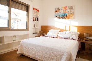 Un dormitorio con una cama grande, cabecera de madera, ventana con persianas, mesita de noche con lámpara, estante con libros y una obra de arte urbana en la pared.
