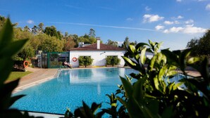 A swimming pool with a clear blue water, surrounded by greenery and a white building with a red roof. - Camping As Cancelas (Santiago de Compostela)