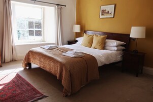 A neatly made bed with a brown bedspread and white pillows, two bedside lamps, a window with curtains, and a framed picture on the wall.
