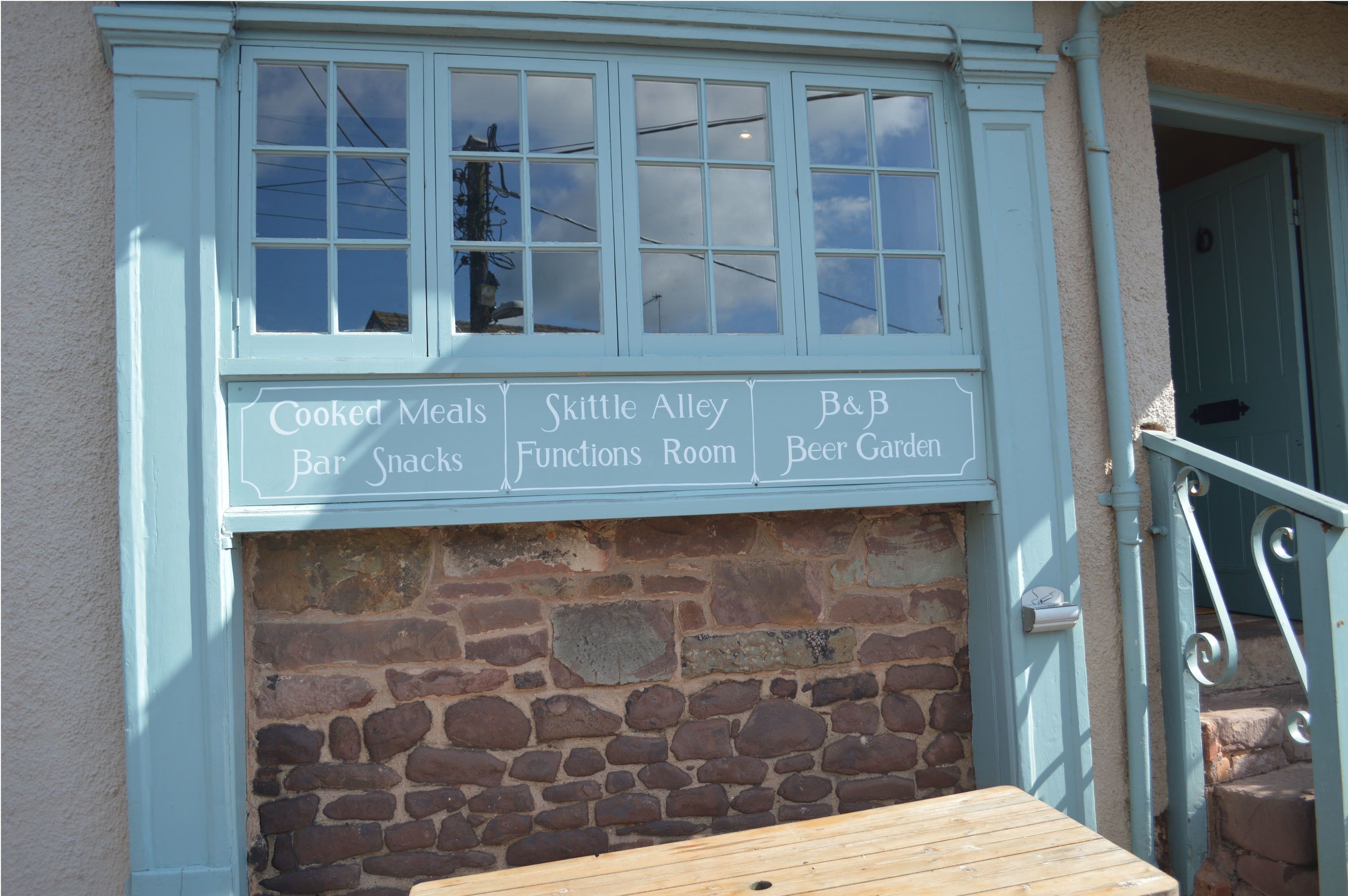 A building with a stone wall and a sign listing amenities such as Cooked Meals, Bar Snacks, Functions Room, and Beer Garden.