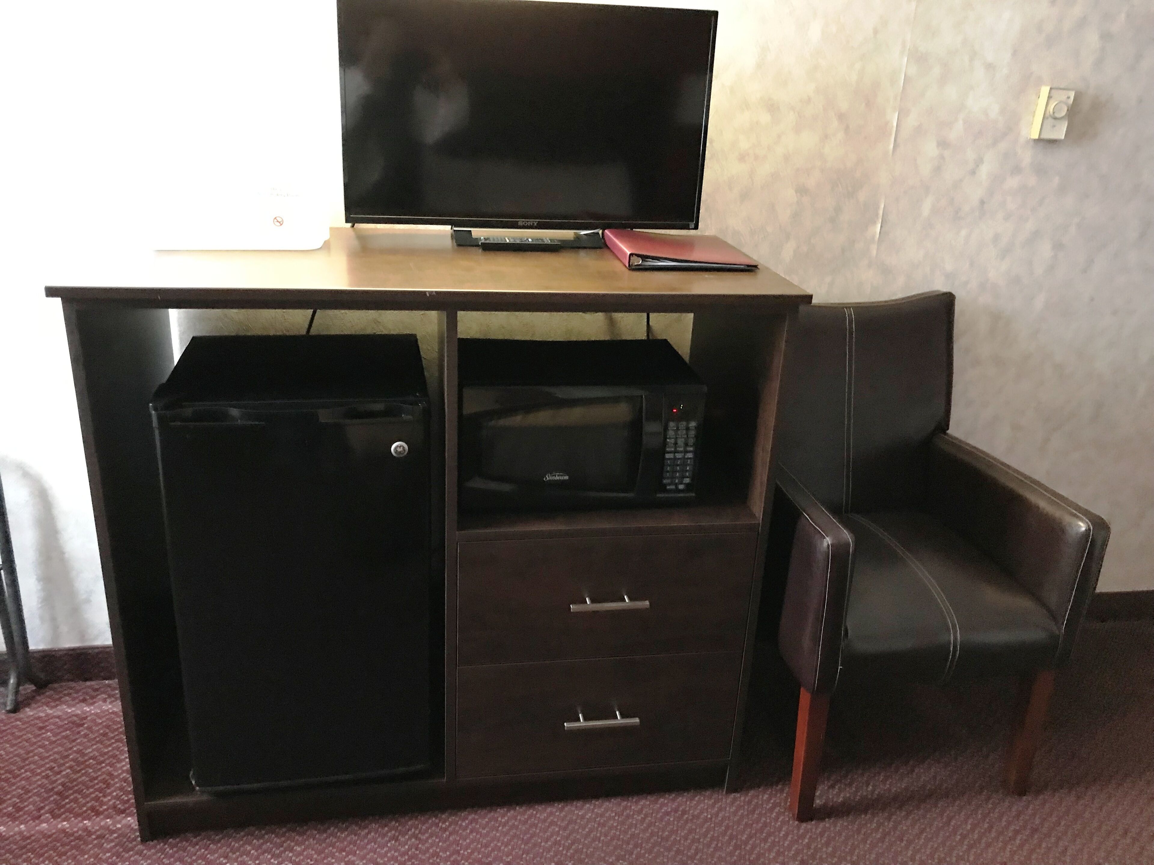 Ett kompakt hotellrum med ett skrivbord i trä, en platt-TV, en mikrovågsugn och ett litet arkivskåp. En läderstol står placerad bredvid skrivbordet.