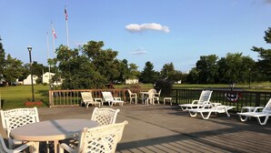 Un espace extérieur aménagé en terrasse, avec des chaises et une table en plastique blanc, entouré d’une plateforme en bois et offrant une vue sur un terrain de golf.