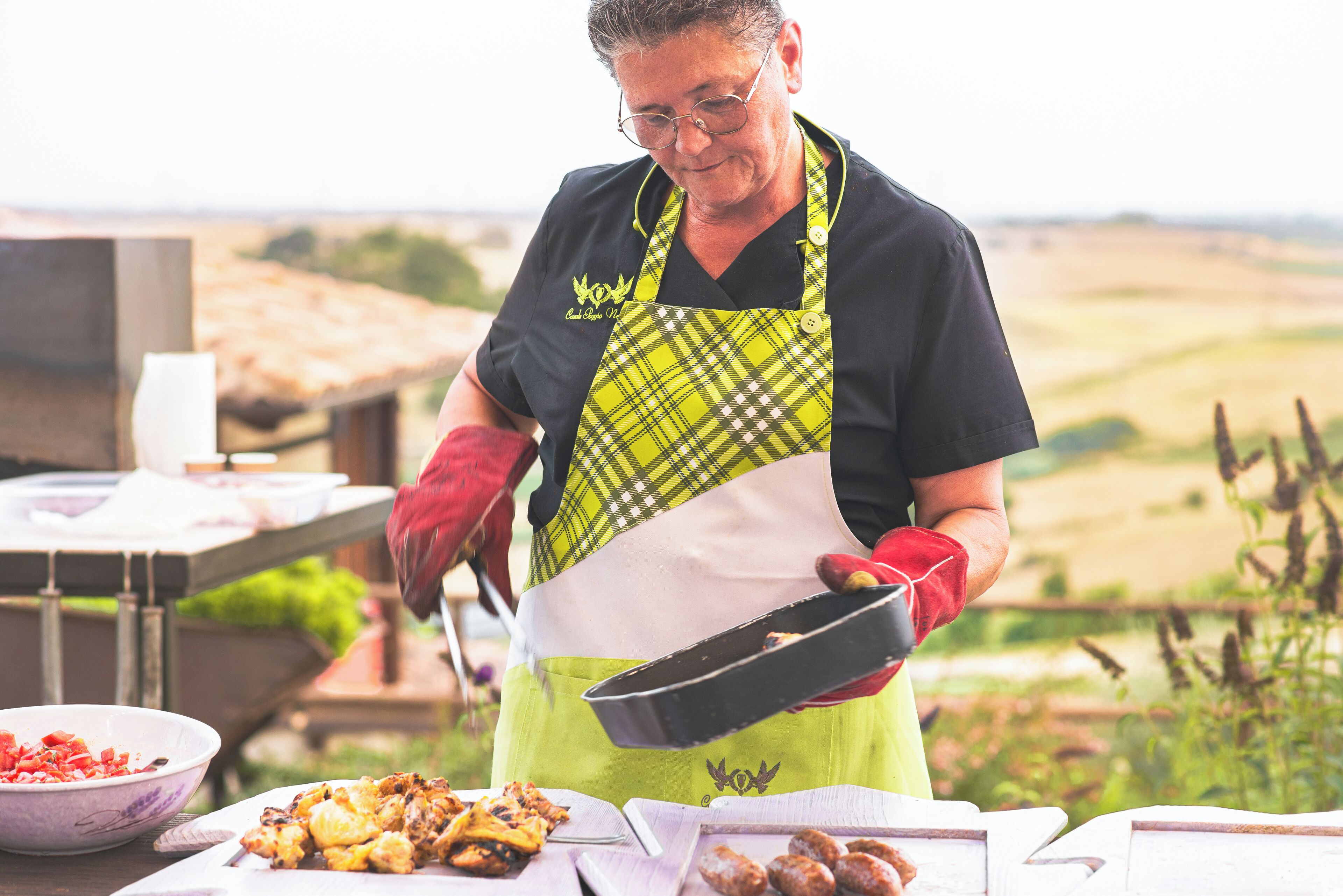 A person in a green apron cooking outdoors with a pan, a bowl of food, and a cutting board with food items.