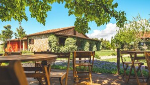 Un espacio de descanso al aire libre de estilo rústico, con mesas y sillas de madera, rodeado de abundante vegetación y un edificio de piedra con techo rojo.