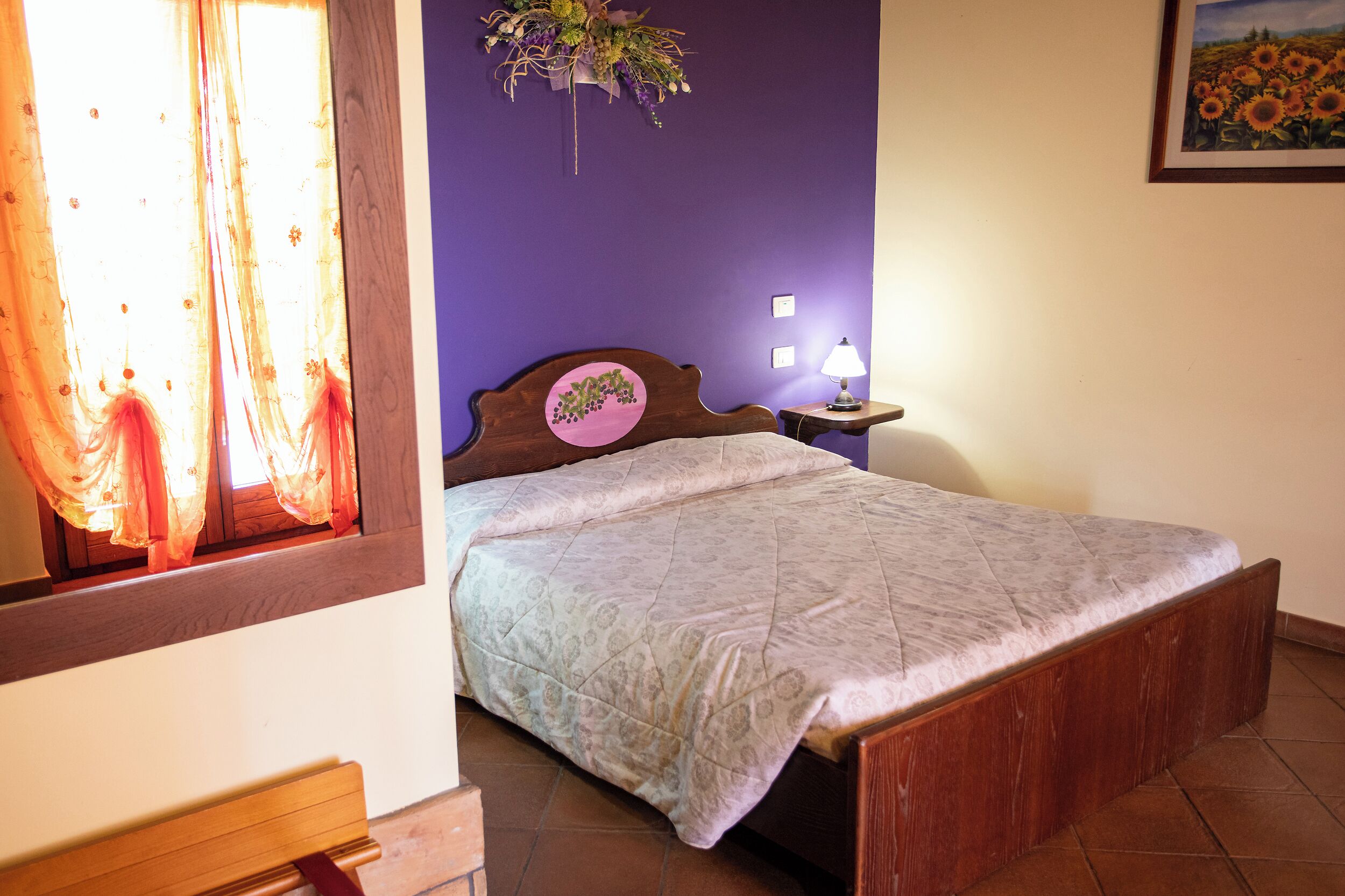 A bedroom with a bed, a nightstand, a wall-mounted lamp, a window with curtains, and a wall painting.