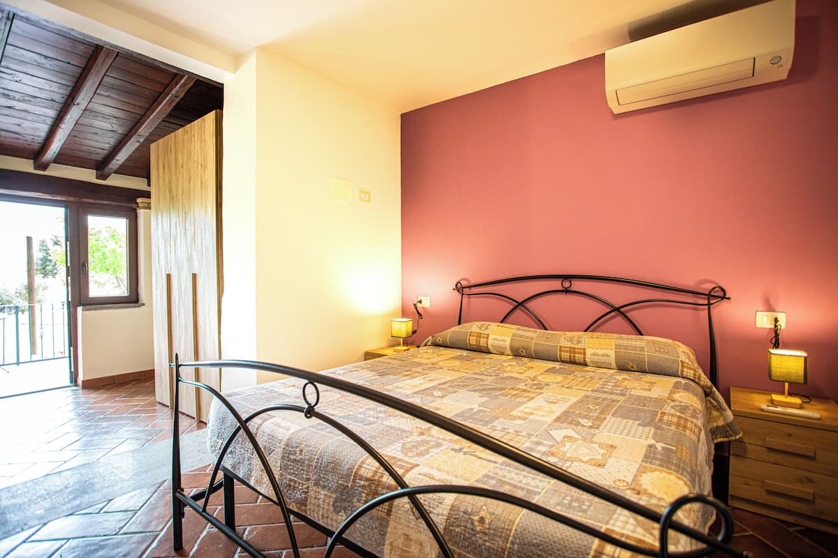 Un dormitorio con estructura de cama metálica, colcha acolchada, mesita de noche de madera con lámpara y pared roja.