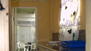 A narrow kitchen with white cabinets, a blue dish rack, and a window with a floral curtain. - Port Lodge (Port of Spain)