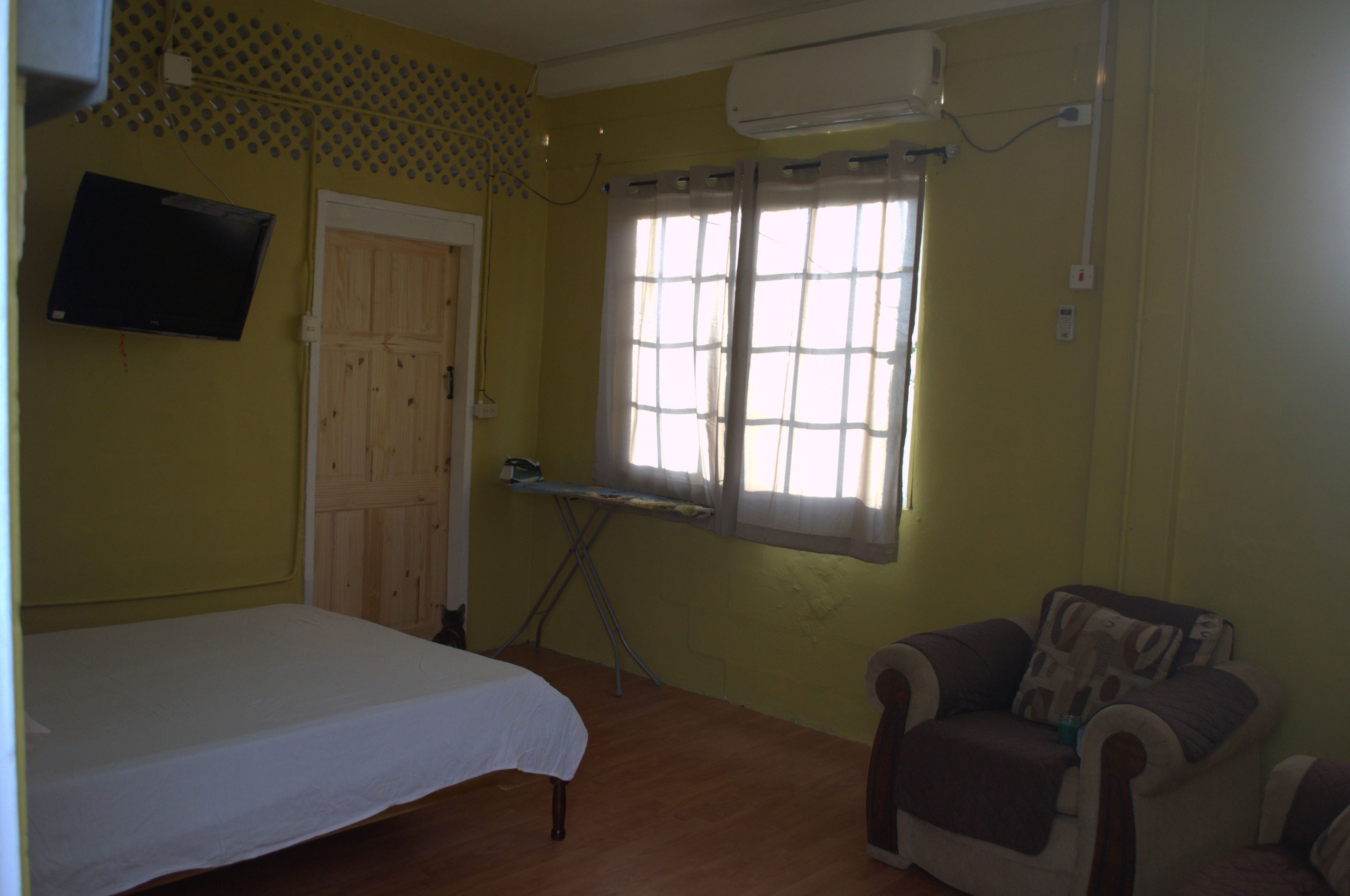 Una habitación con cama, una silla, una ventana con cortinas, una tabla de planchar y un televisor colocado en la pared.