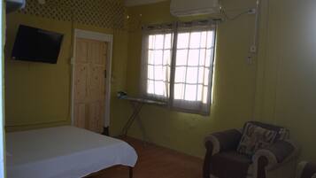 Una habitación con cama, una silla, una ventana con cortinas, una tabla de planchar y un televisor colocado en la pared.