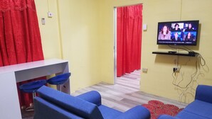 A room with a TV displaying a news broadcast, a white desk with two blue chairs, and a red curtain. - Port Lodge (Port of Spain)
