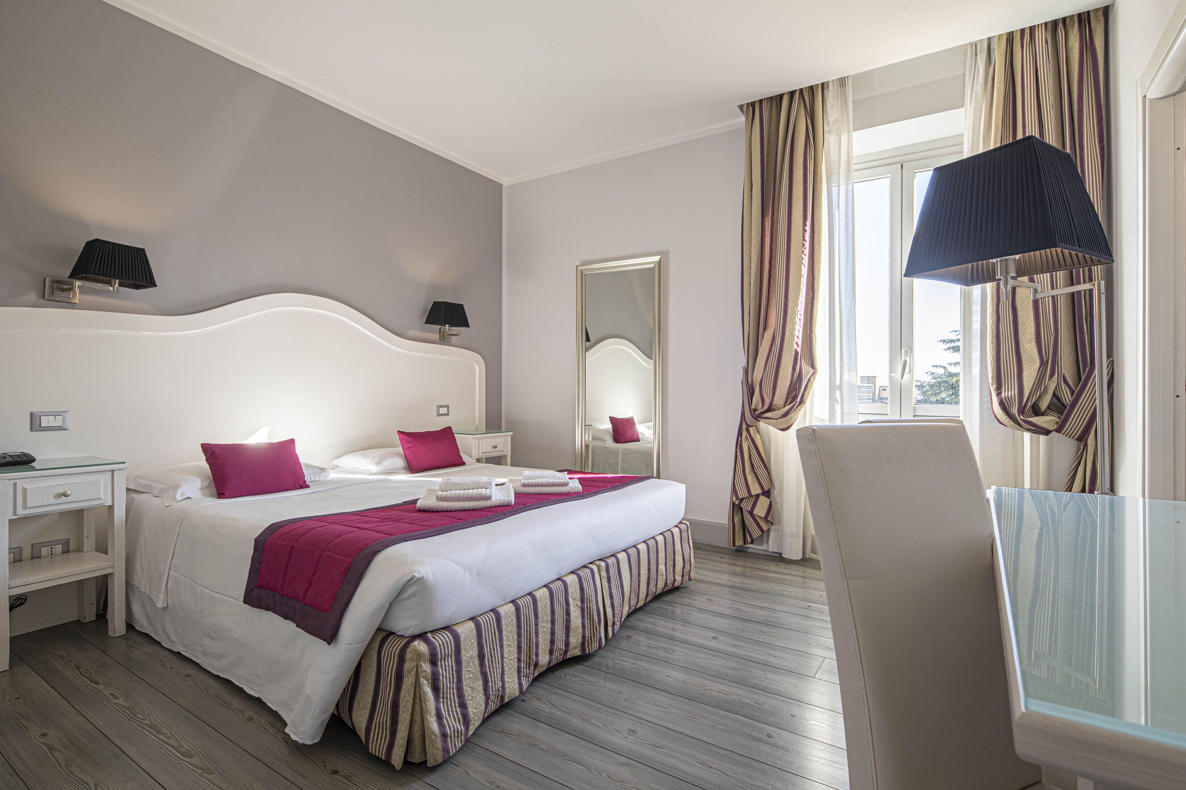 Une chambre d’hôtel avec un grand lit, une table de chevet, un miroir, une chaise et une fenêtre avec des rideaux.