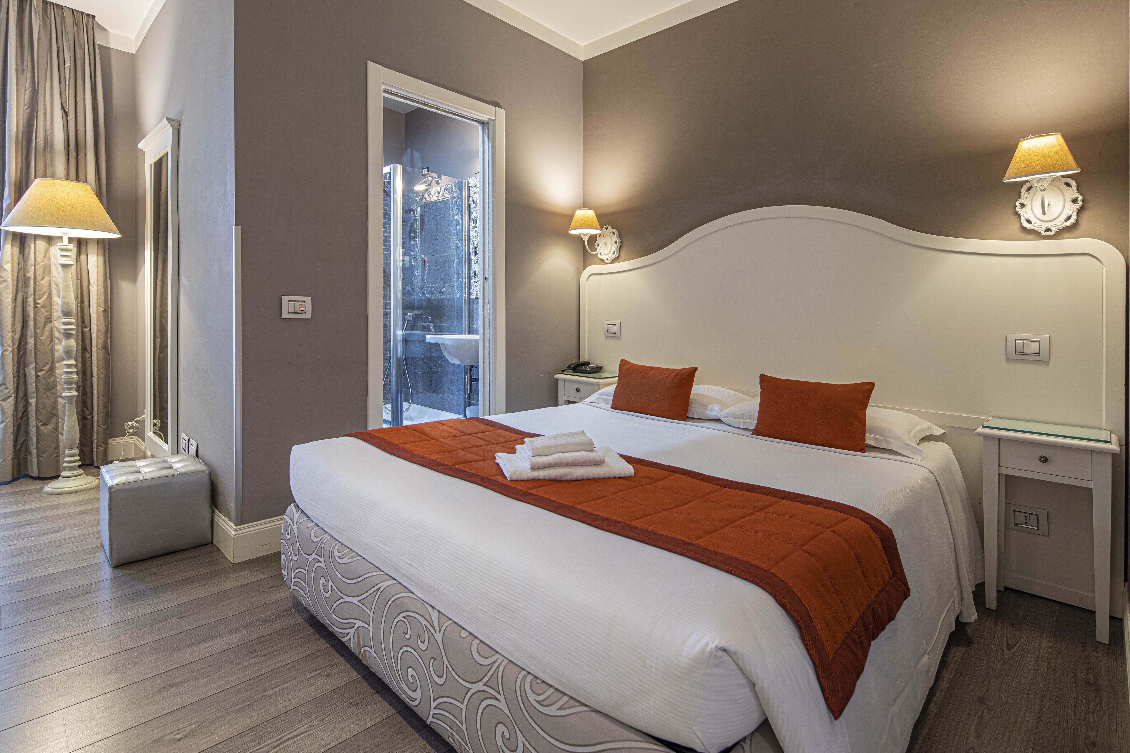 Quarto de hotel com uma cama grande, duas mesas de cabeceira, um criado-mudo, porta de box de vidro e uma luminária de cada lado da cama.
