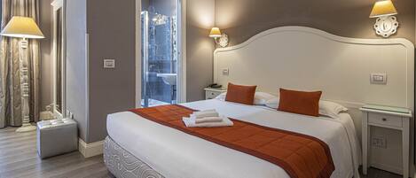 A hotel room with a large bed, two bedside tables, a nightstand, a glass shower door, and a lamp on each side of the bed.